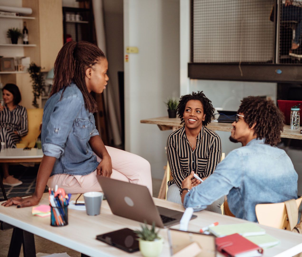 Three people have a relaxed conversation in a modern office, with one woman sitting on a desk and two others seated nearby. Laptops, notebooks, and office supplies are visible on the desk around them.