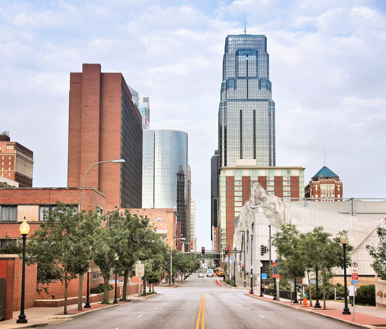 A city street flanked by trees and modern buildings, leading toward tall skyscrapers under a partly cloudy sky. The street is empty, creating a calm urban scene.