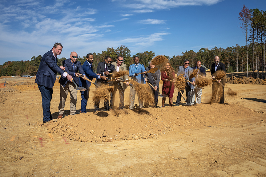 A group of people in business attire participate in a groundbreaking ceremony, using shovels to toss dirt into the air at a construction site under a clear blue sky.