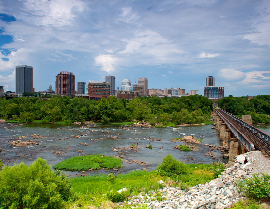 A city skyline with modern buildings rises behind a river with rocks and lush green trees. A railroad bridge crosses the river from the right side of the image under a partly cloudy sky.