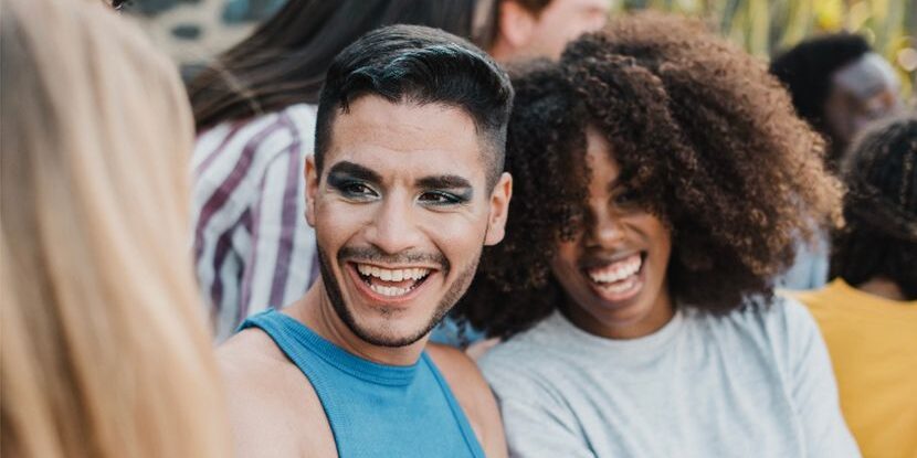 A group of friends sits together outdoors, smiling and laughing. In the foreground, a person with short dark hair, and a blue top is next to a woman with curly hair in a gray shirt.