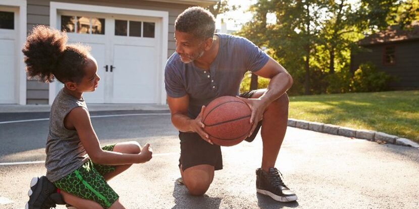 A man kneels on a driveway holding a basketball, smiling and talking with a young girl who is sitting on one knee. They appear to be playing or practicing basketball outside on a sunny day.
